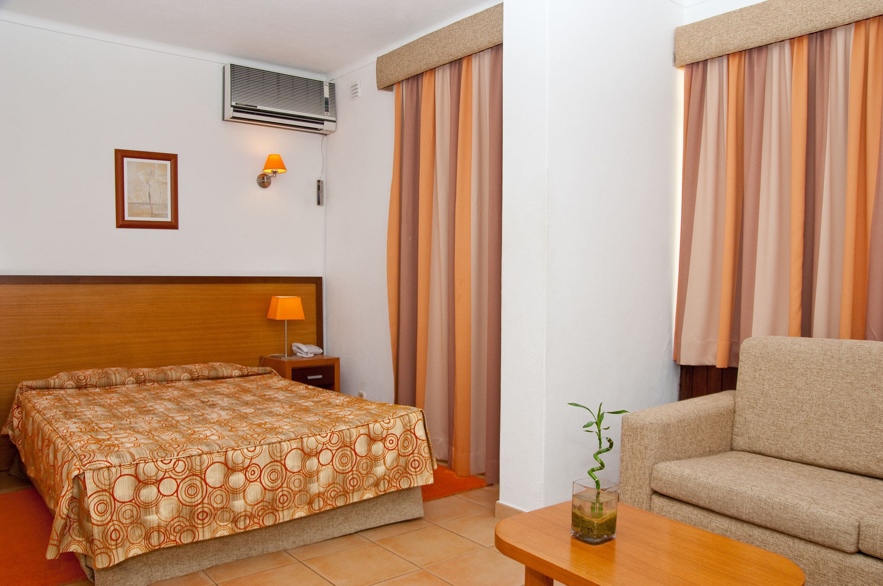 Een hotelkamer met een bed, een slaapbank, een nachtkastje, een lamp, een airconditioner en gordijnen.