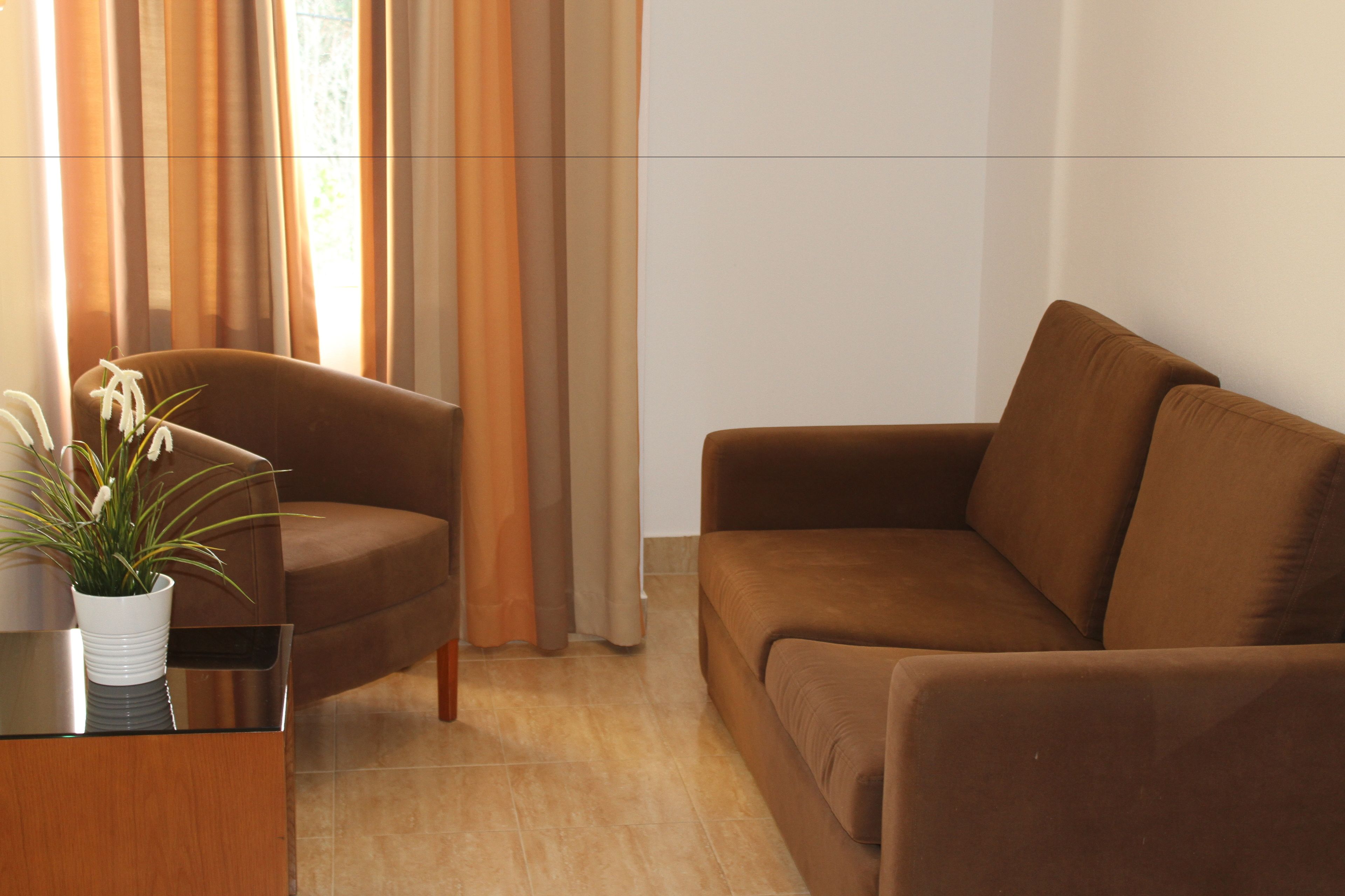 Ein modernes Wohnzimmer mit einer braunen Couch, einem braunen Sessel und einem Glastisch mit weißer Vase und Pflanze.