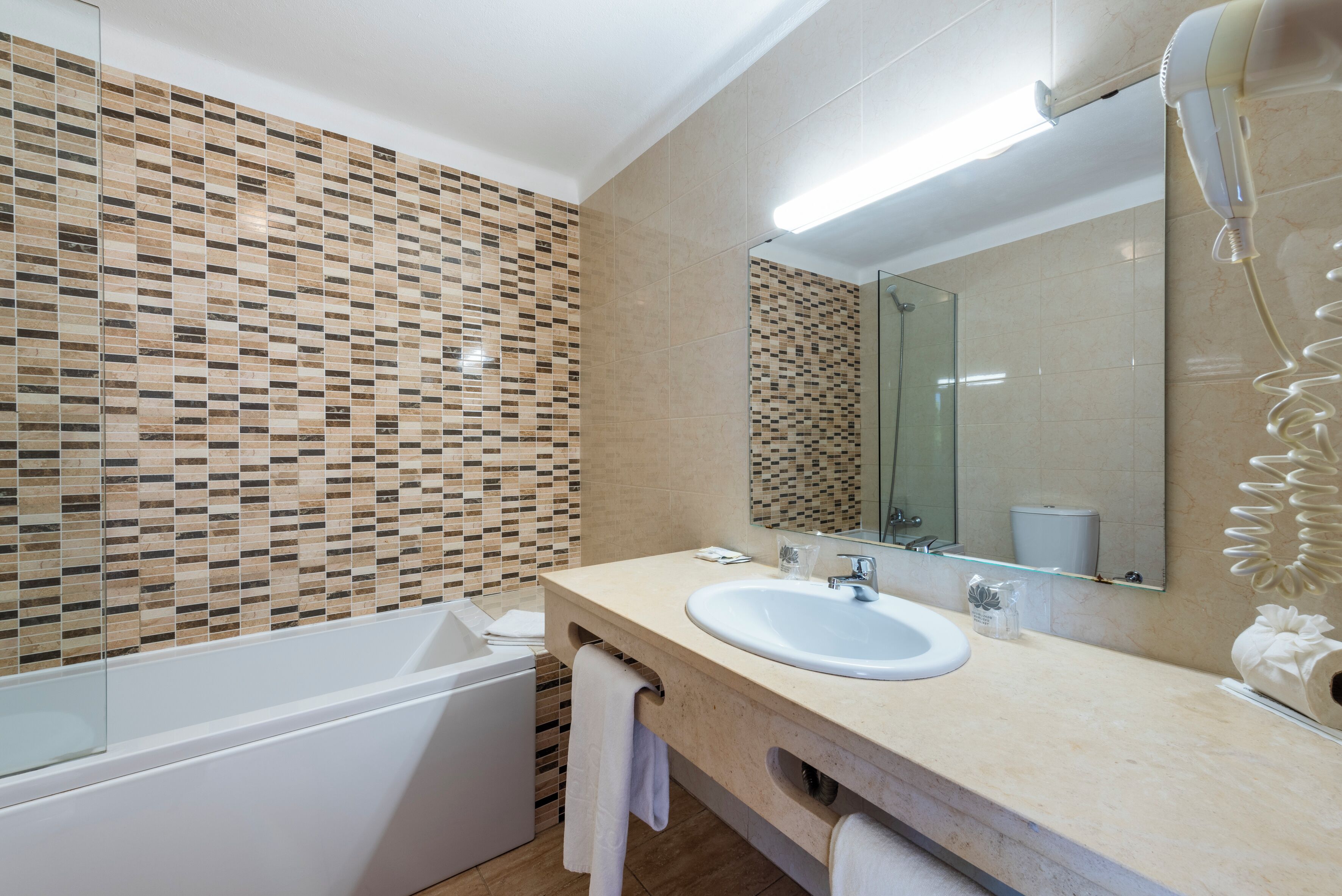 A modern bathroom with a mosaic tile wall, a white bathtub, a sink with a mirror, and a hairdryer on the counter.