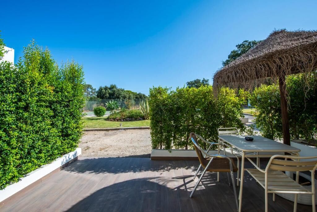 Un patio al aire libre con terraza de madera, mesa y sillas, un parasol de paja y abundante vegetación.