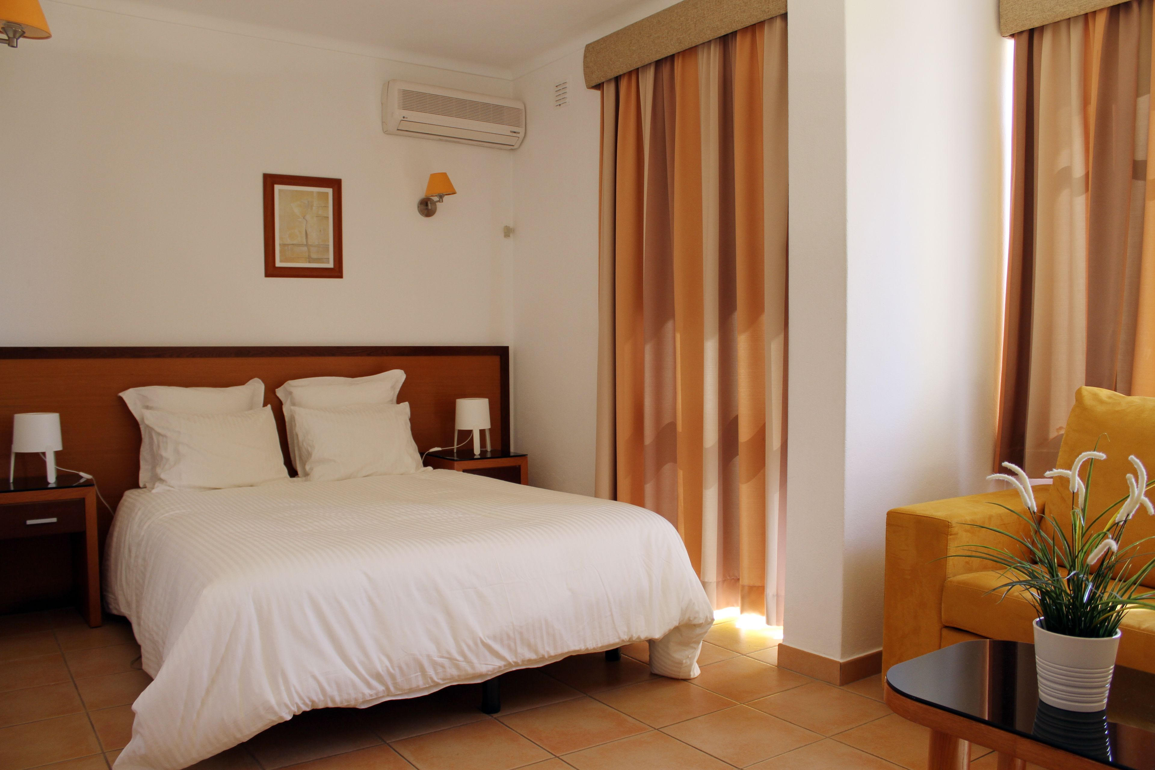 Une chambre d’hôtel avec un grand lit, deux tables de chevet avec des lampes, une petite table avec une plante et une chaise.