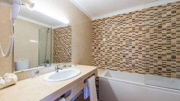 Une salle de bain avec un comptoir beige, un lavabo blanc, une douche en verre et un mur recouvert de mosaïques.