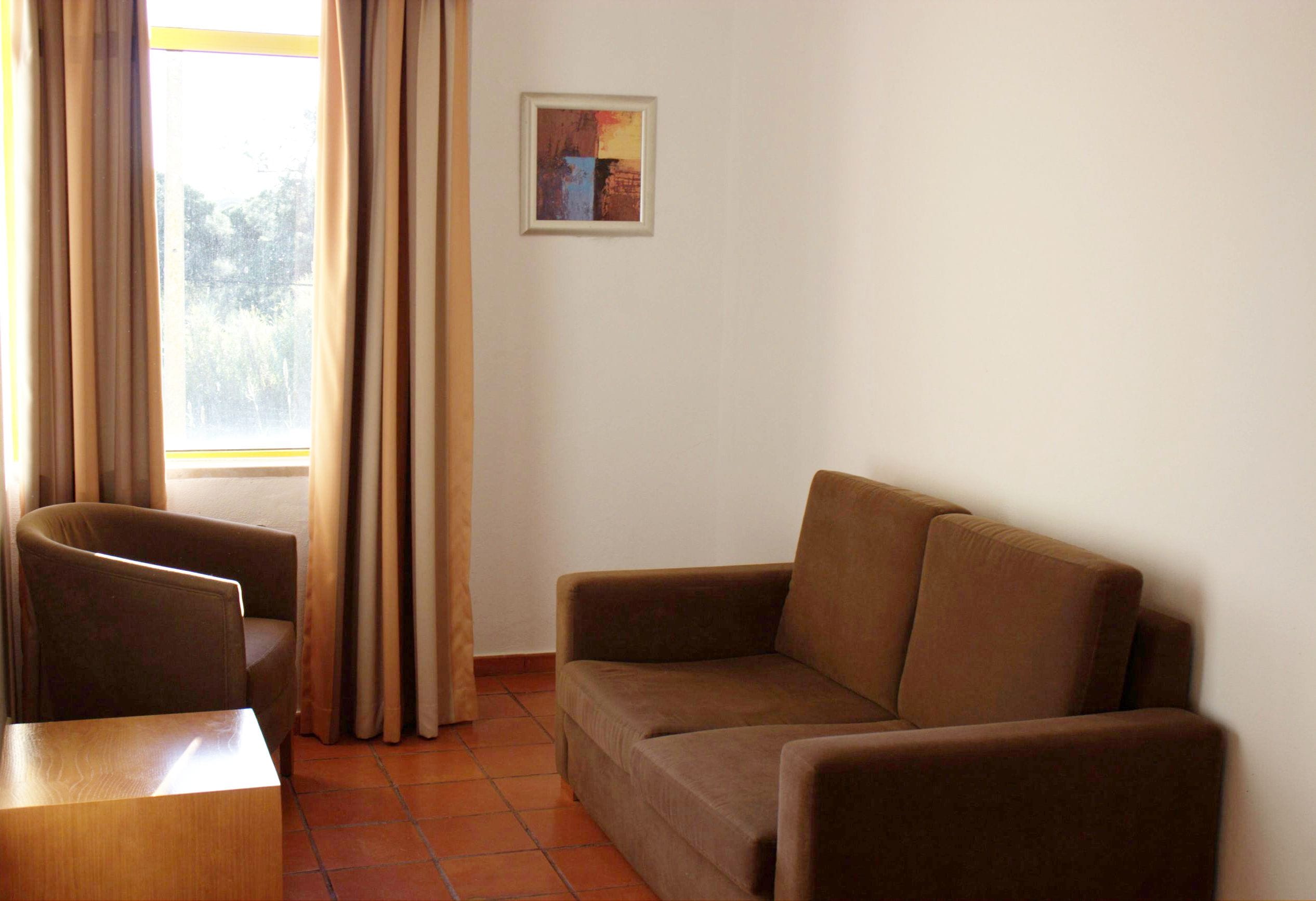 A room with a brown sofa, a chair, a small table, and a window with curtains.