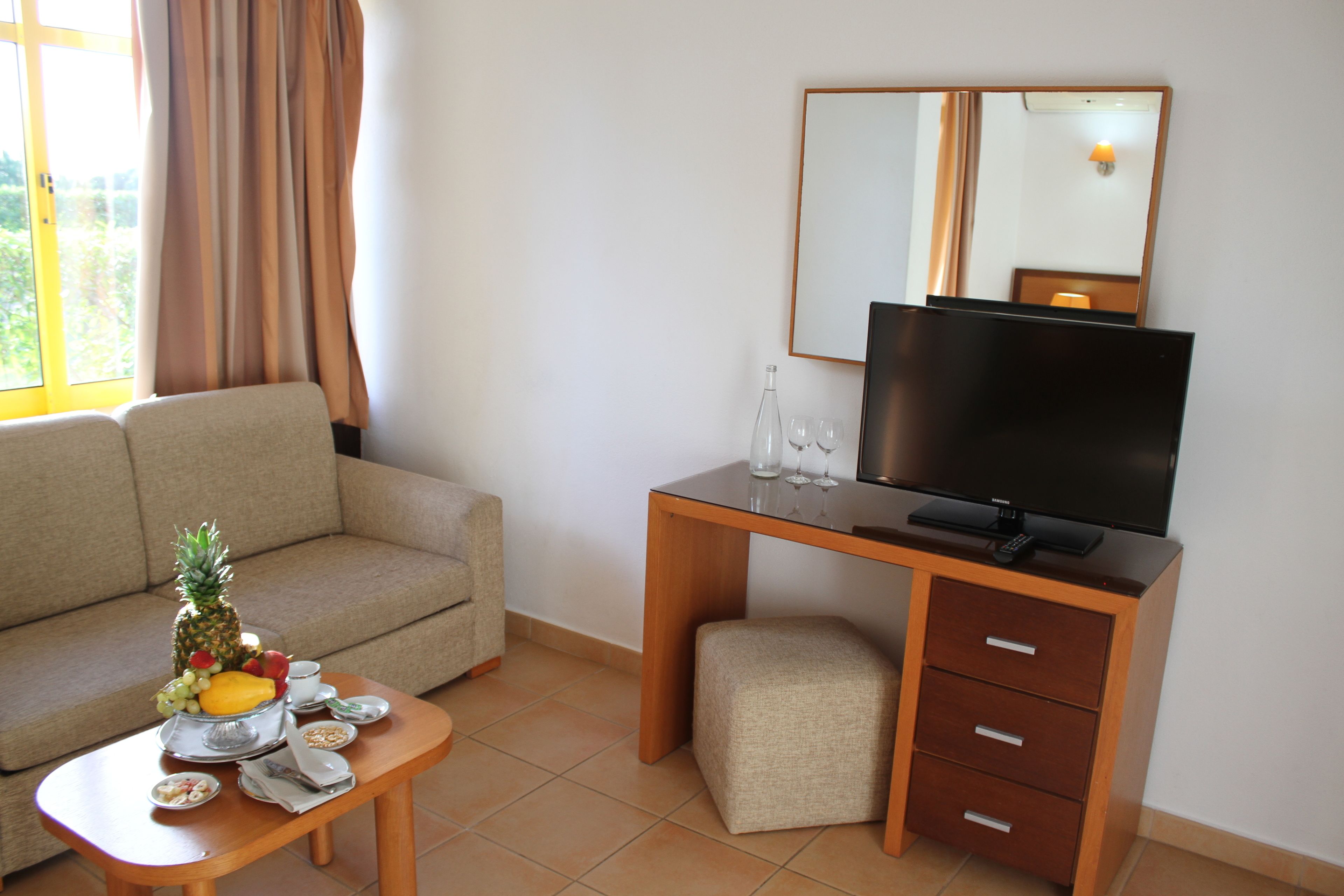 Quarto de hotel com sofá, escrivaninha de madeira com televisão, um banquinho pequeno e mesa com frutas e copos.