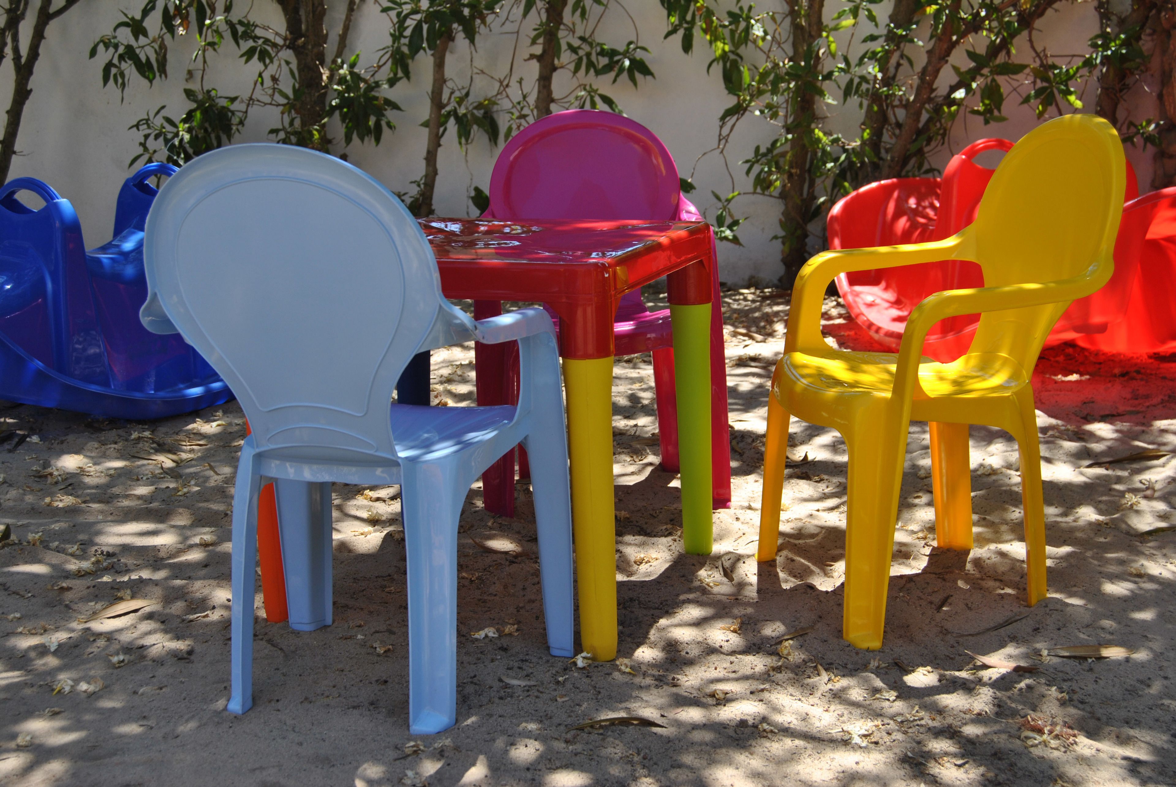 A set of colorful plastic chairs and tables arranged outdoors on sandy ground, with a wall and foliage in the background.
