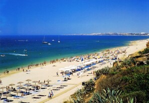 A beach with sun loungers and umbrellas, a clear blue sea, and boats in the distance. - Hotel Pinhal do Sol (Loulé)