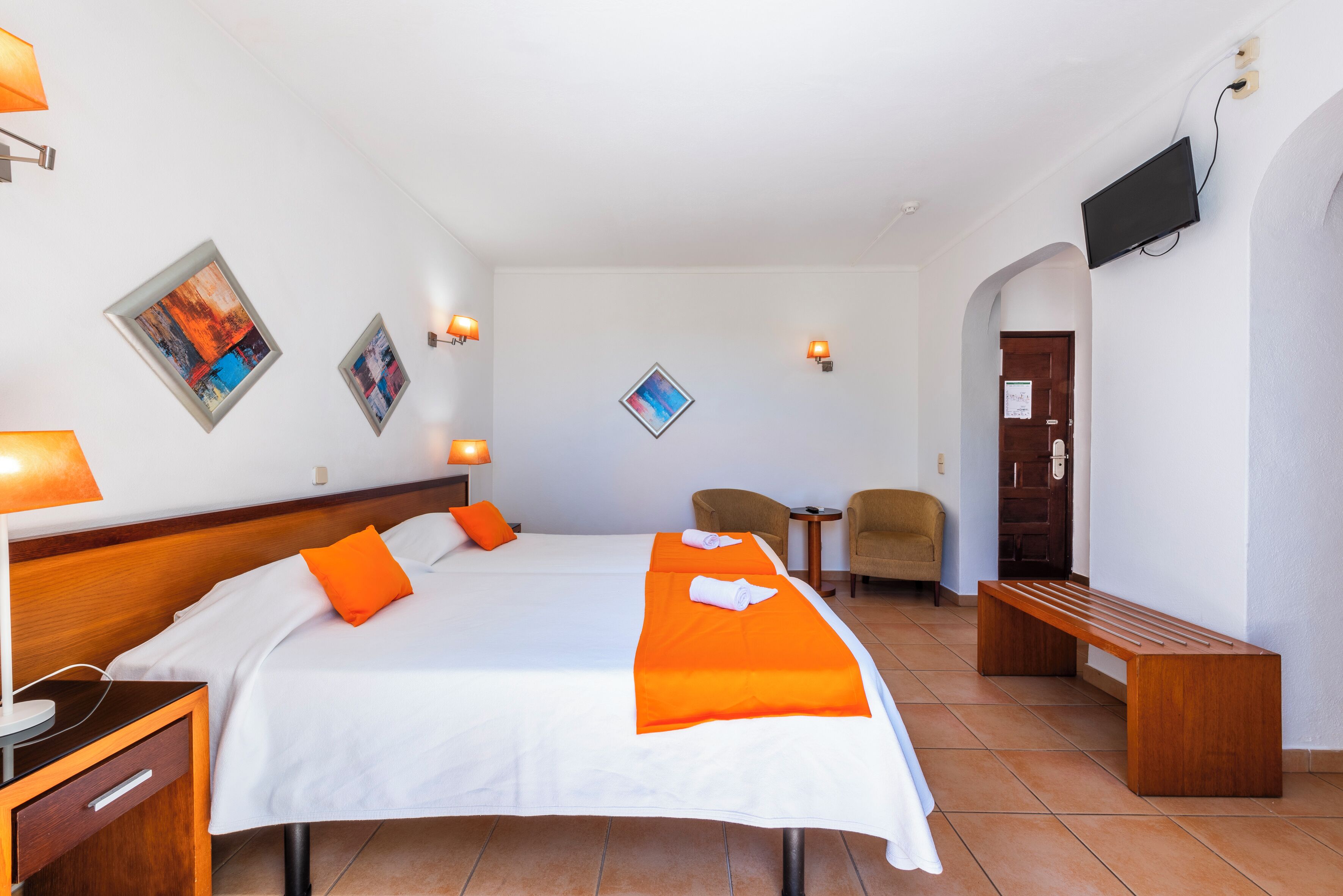Une chambre d’hôtel avec deux lits, un bureau, une chaise, un banc et des œuvres d’art accrochées aux murs.