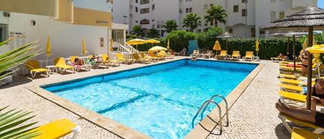 Una piscina con sillas de descanso amarillas y sombrillas, rodeada por un patio empedrado y un edificio con balcones.