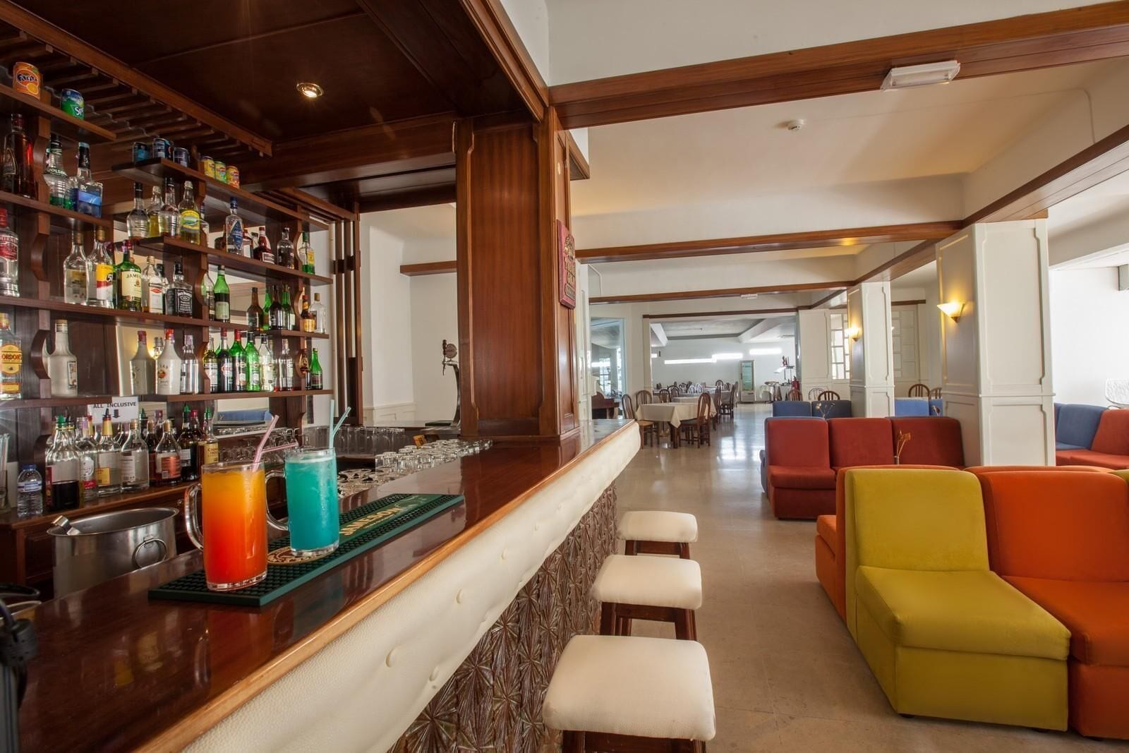 Een bar met verschillende drankflessen, een bargedeelte en een zithoek met kleurrijke banken.
