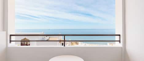 A balcony with a white table and chairs, overlooking a coastal view with buildings and the sea.