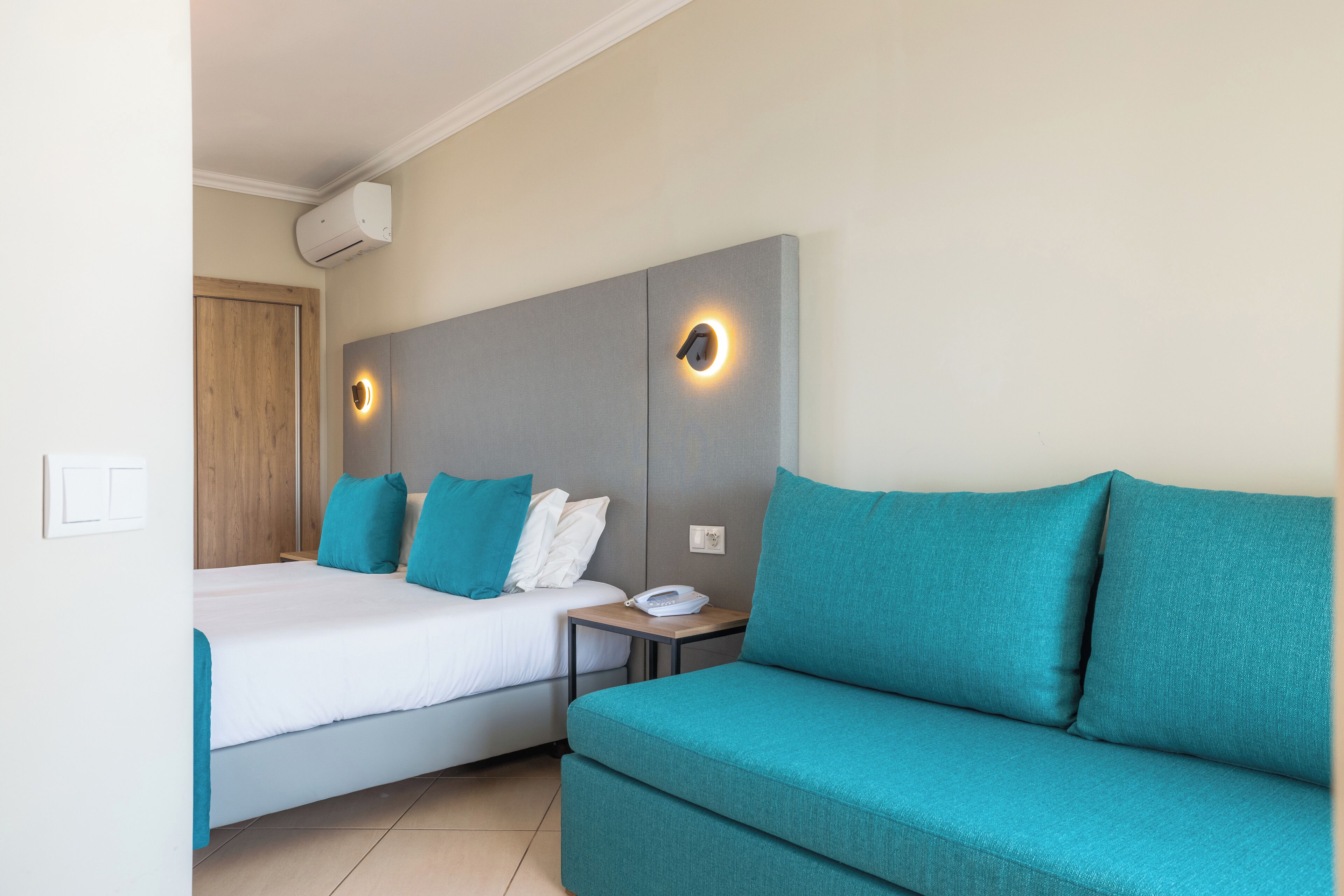 Habitación de hotel con cama, mesita de noche con teléfono, sofá color turquesa y lámpara fijada a la pared.