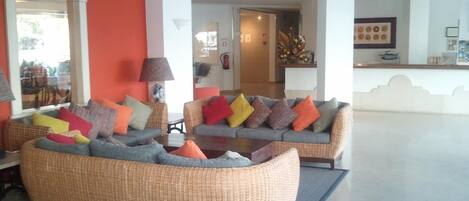 A spacious lobby with two grey sofas adorned with colorful cushions, a wicker armchair, and a coffee table.