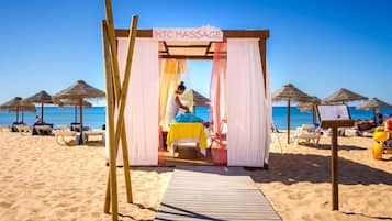 Una carpa para masajes en la playa con estructura de madera y cortinas blancas, ubicada en una playa arenosa con sombrillas y sillas reclinables.