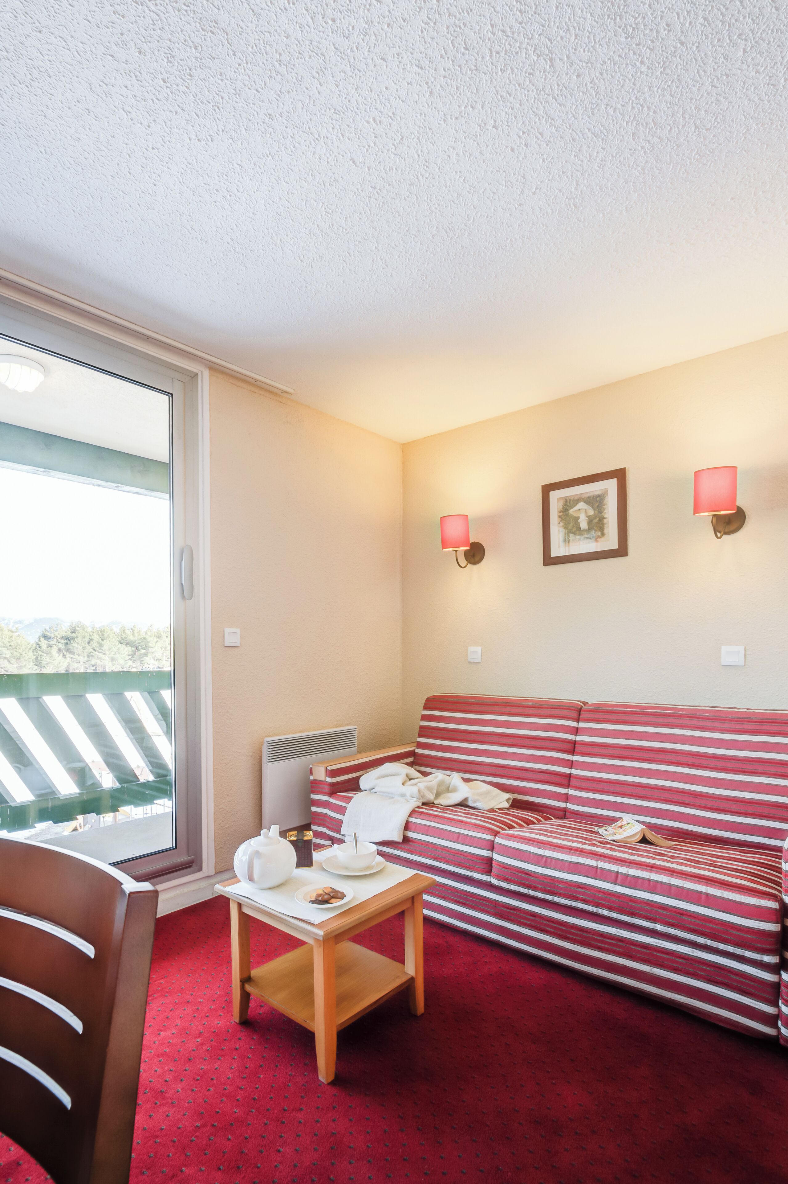 A cozy room with a striped sofa, a small table with a teapot and cups, a chair, and a view through a sliding door.