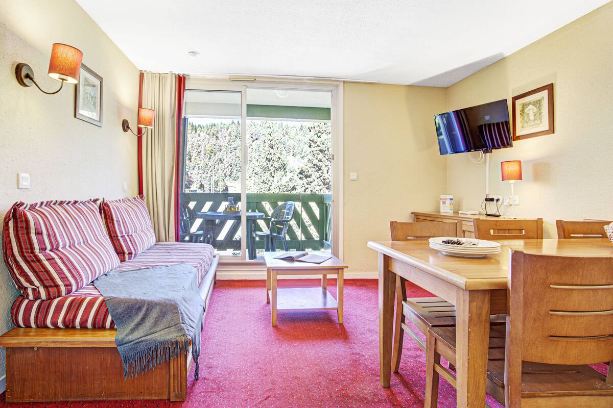 A cozy room with a sofa, a dining table, and chairs. There is a large window with a view of trees.