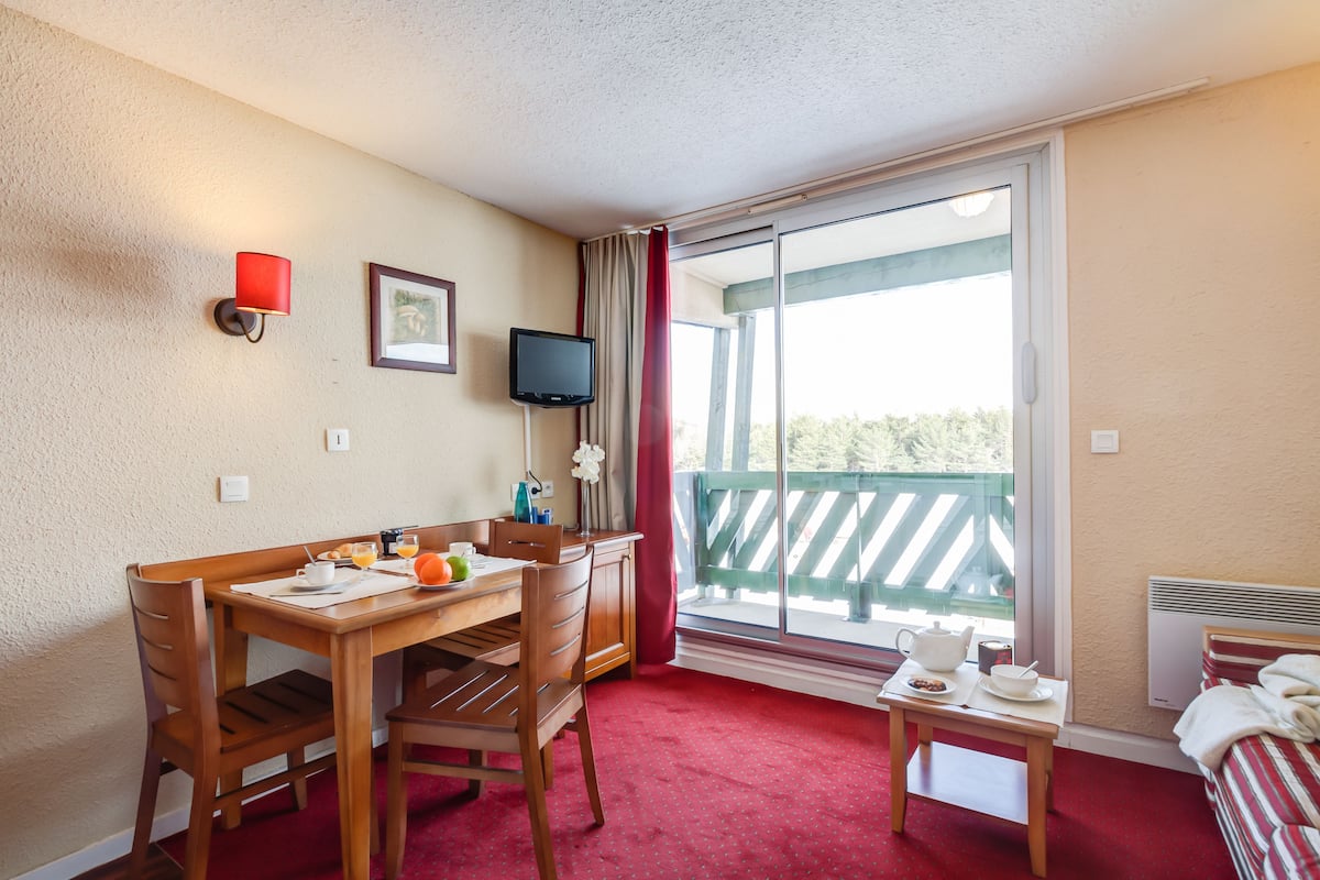 A hotel room with a wooden desk, chairs, a television, a balcony with a view, and a bed with striped bedding.