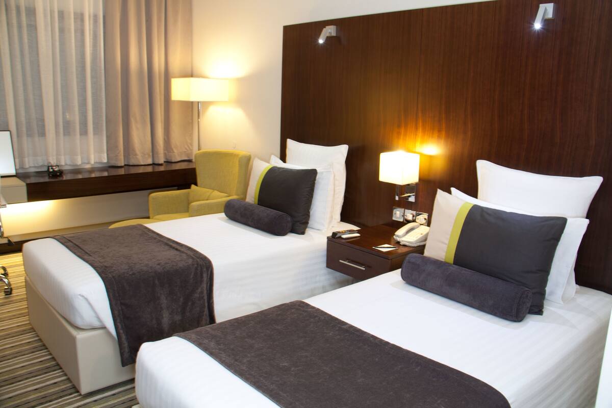 A hotel room with two beds, a desk, a chair, and a nightstand with a lamp.