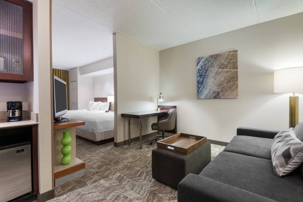 Quarto de hotel com cama, escrivaninha, sofá e pequena cozinha.
