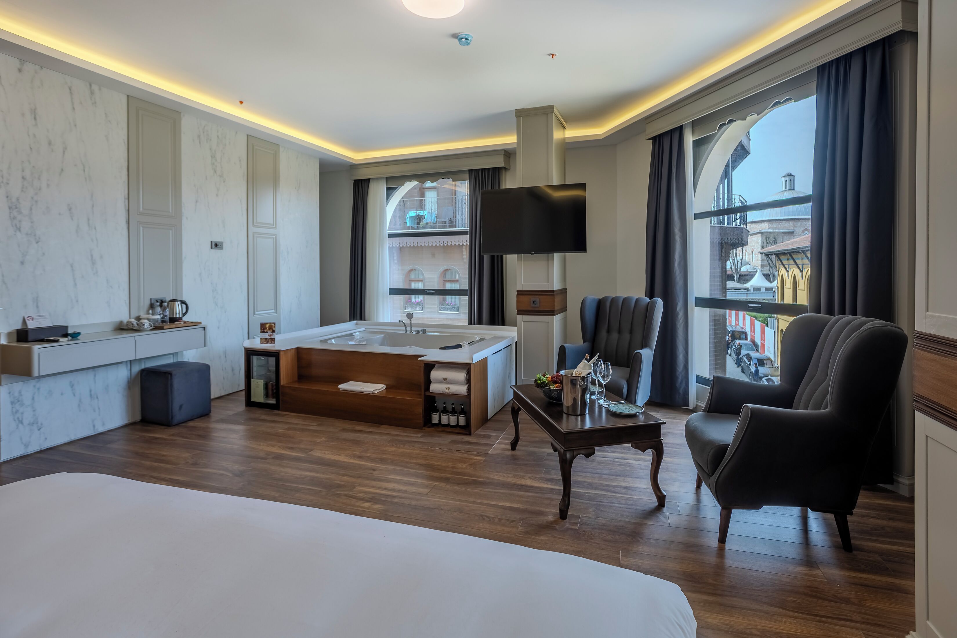 A hotel room with a large bed, a sitting area with a table and chairs, a bathtub, a TV, and a view of the city.