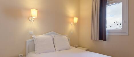 Habitación de hotel con cama, dos mesitas de noche, dos lámparas de pared, una ventana con cortinas y vista a un paisaje nevado.