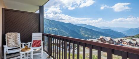 A balcony with two white chairs, a small table with a tray, and a view of mountains and a village.
