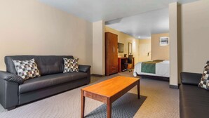 Une chambre d’hôtel avec un canapé en cuir foncé, une table basse en bois, un lit recouvert d’une couverture verte, un bureau avec une chaise et une armoire.