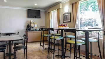 A dining area with a TV mounted on the wall, a window with curtains, and a bowl of fruit on the counter.