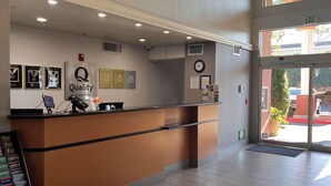 Un espace d’accueil d’hôtel comprenant un comptoir de réception, des trophées encadrés, une horloge et une vue sur l’extérieur à travers la porte vitrée.