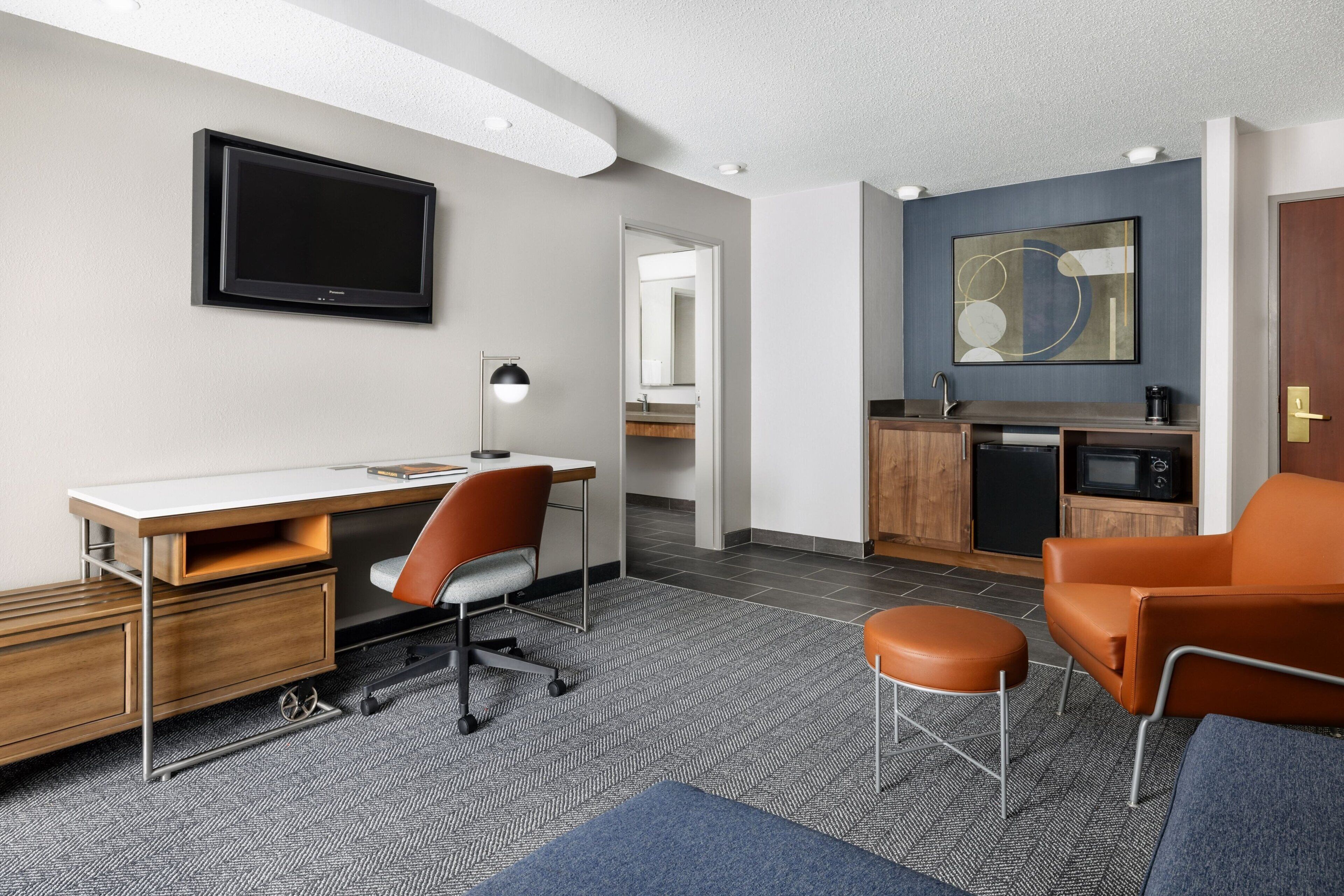 A modern hotel room with a desk, chair, and seating area. It includes a television, kitchenette, and a large mirror.