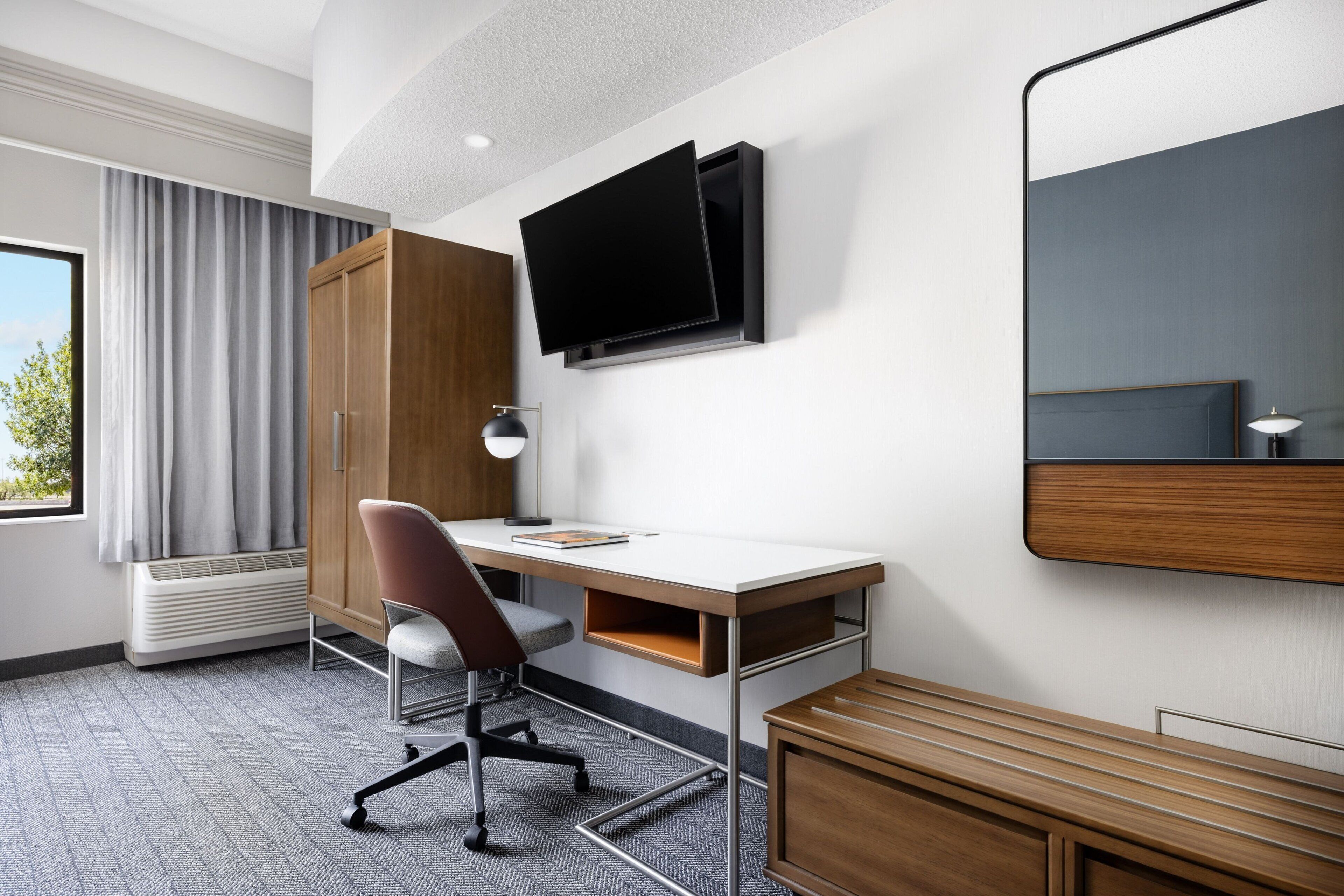 Habitación de hotel con escritorio, silla, televisor, armario y un espejo grande.