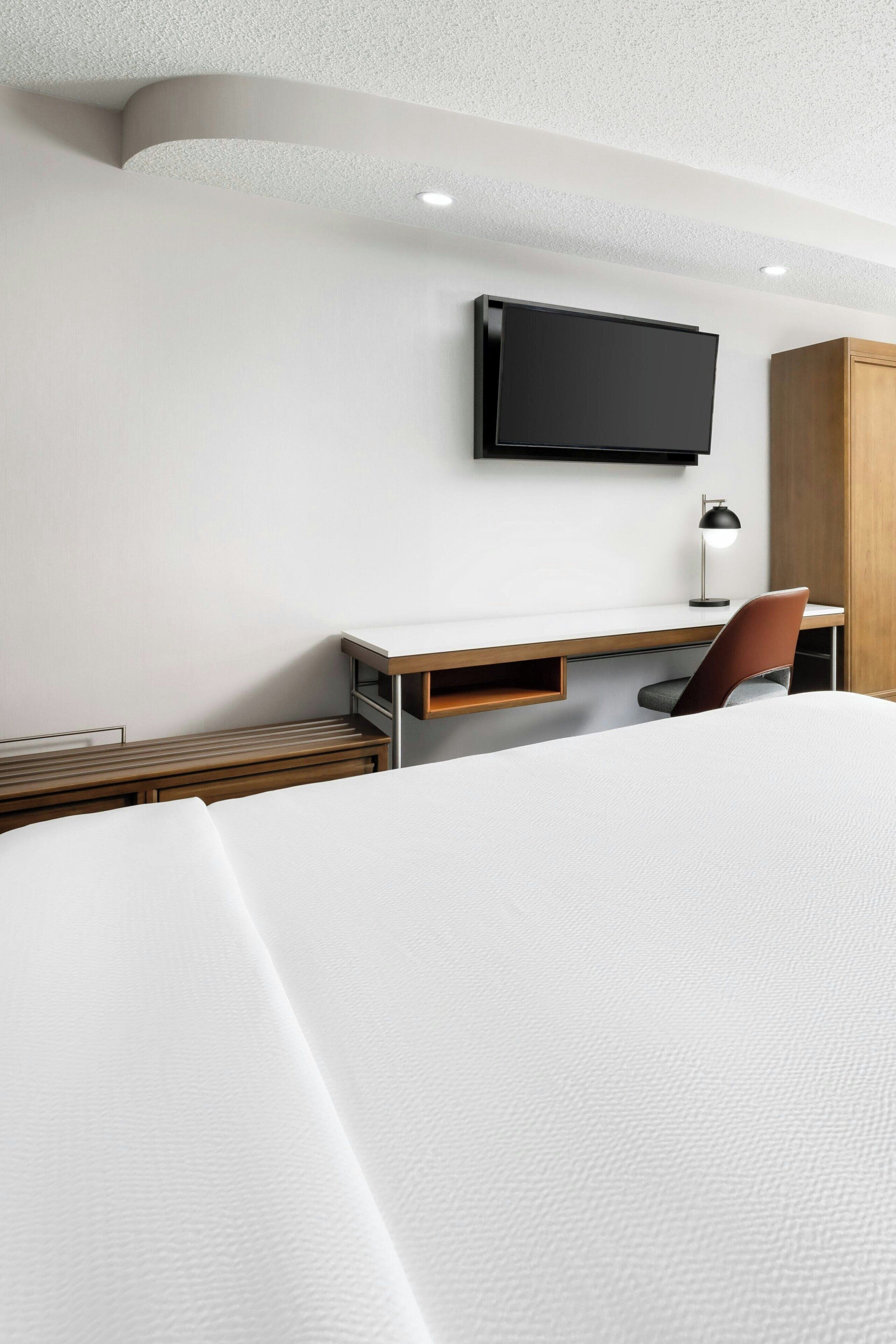 Habitación de hotel con cama, escritorio con silla, televisor montado en la pared y armario de madera.