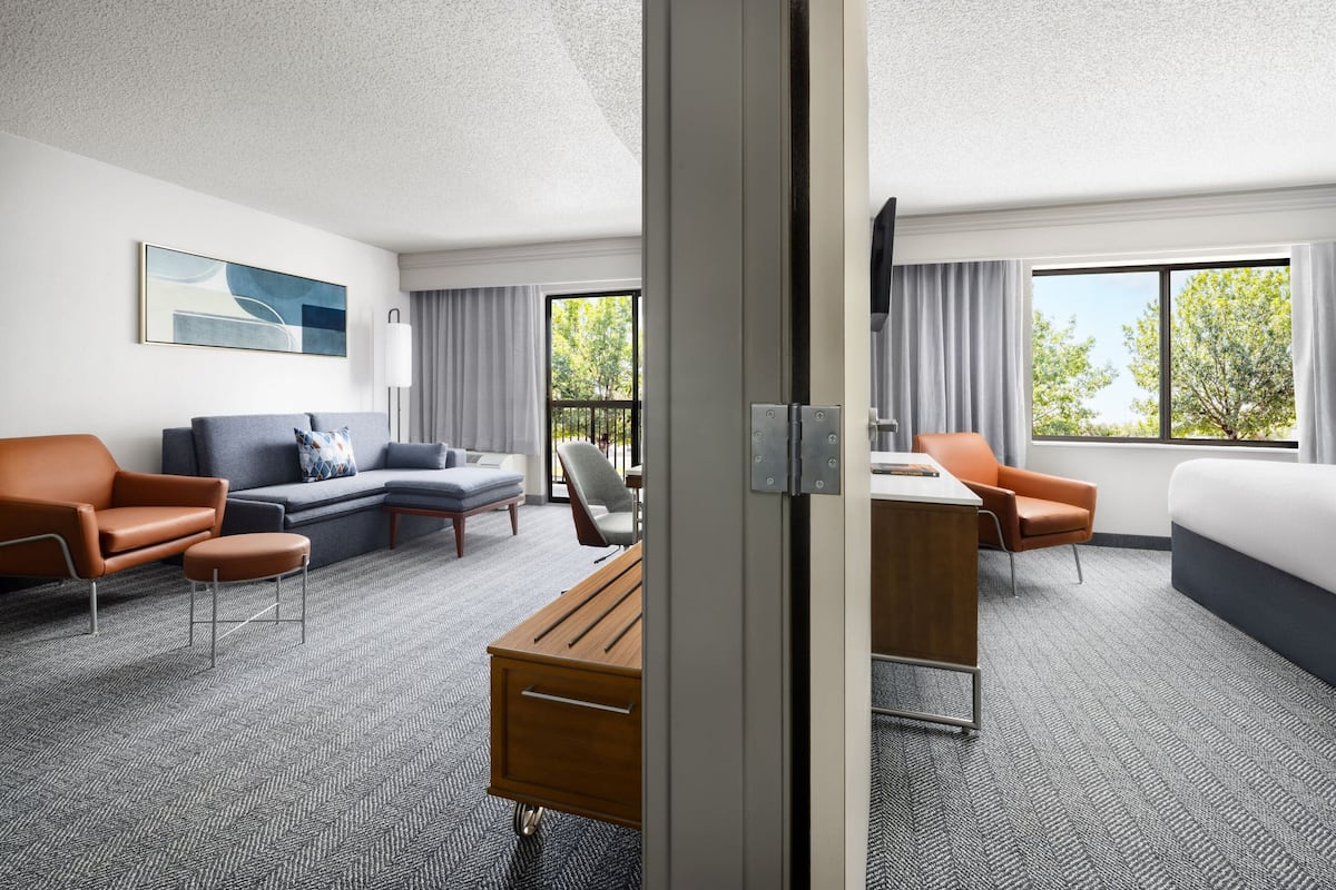 Ein Hotelzimmer mit Sofa, Ohrensessel, Schreibtisch und Blick auf Bäume durch große Fenster.