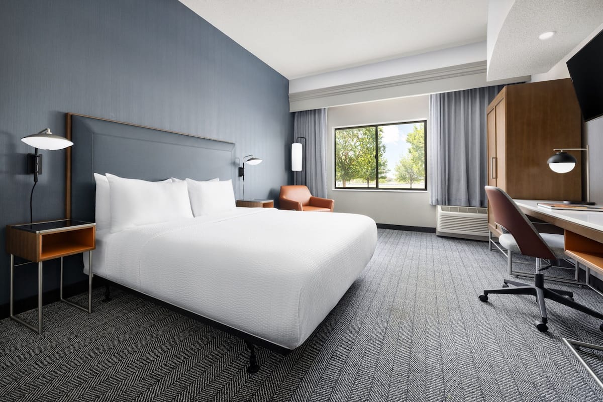 Ein Hotelzimmer mit einem großen Bett, einem Schreibtisch, einem Sessel, einer Lampe und einem Fenster mit Vorhängen.