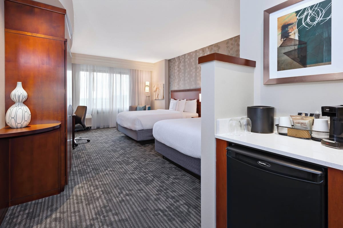 Ett hotellrum med två sängar, ett skrivbord med en kaffebryggare, en lampa och ett inramat konstverk på väggen.
