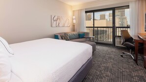 Habitación de hotel con una cama grande, un sofá, un escritorio y vistas a la ciudad desde un balcón.