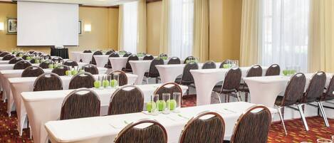 A conference room with multiple tables and chairs, a projector screen, and large windows with curtains.