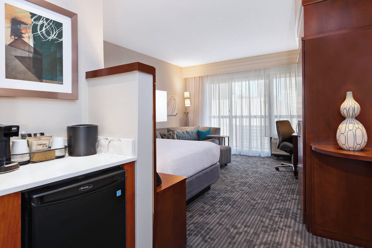 Ett hotellrum med en säng, ett skrivbord och en stol, ett litet pentry och ett stort fönster med gardiner.