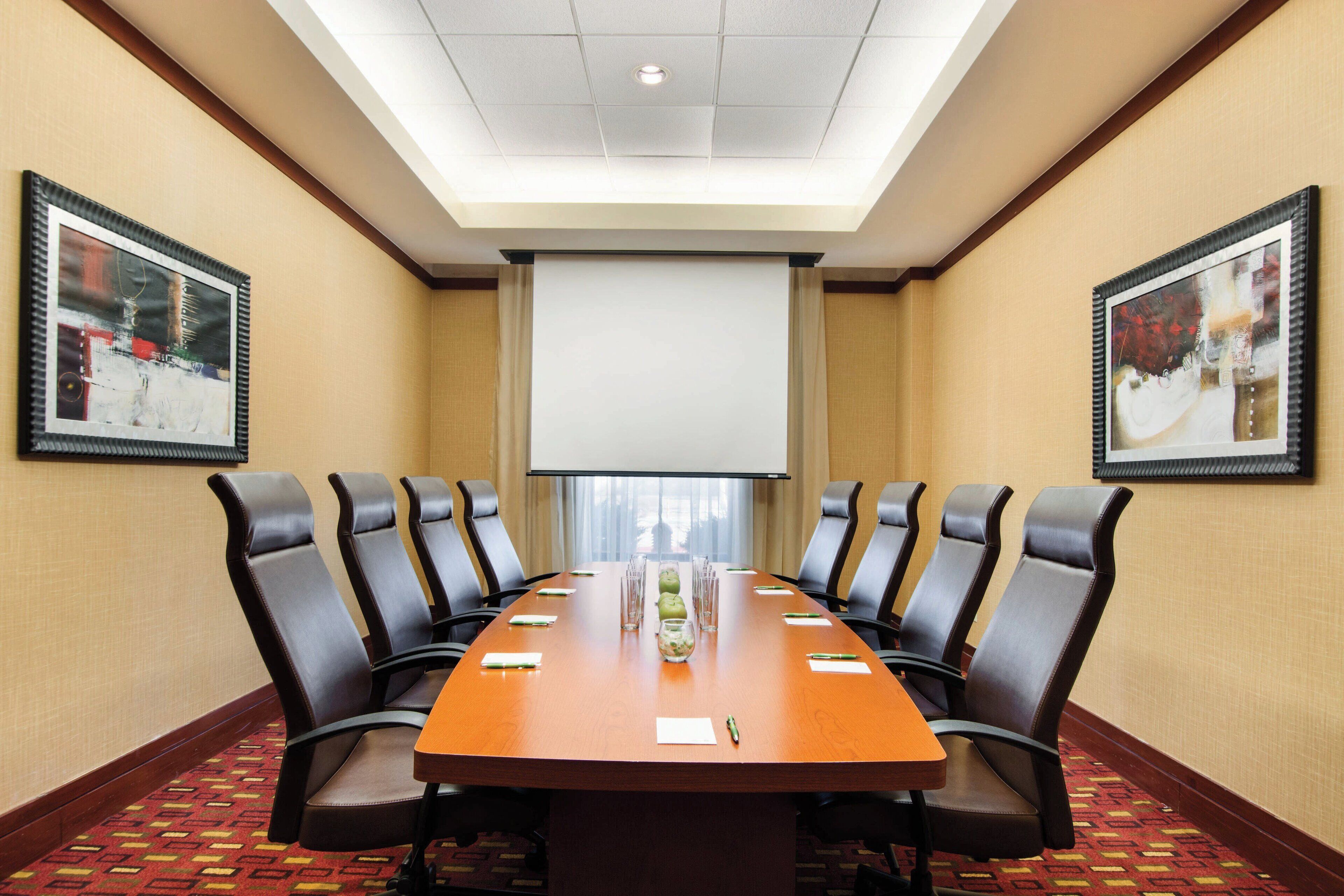 A conference room with a long table, 10 chairs, a projector screen, and two framed pictures on the walls.