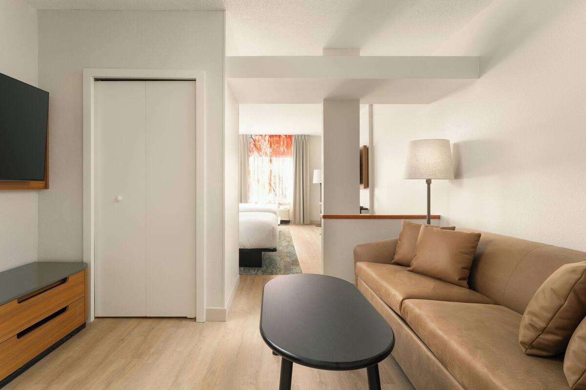 Habitación de hotel moderna con sofá, una mesita, televisor y puerta que da a un dormitorio.