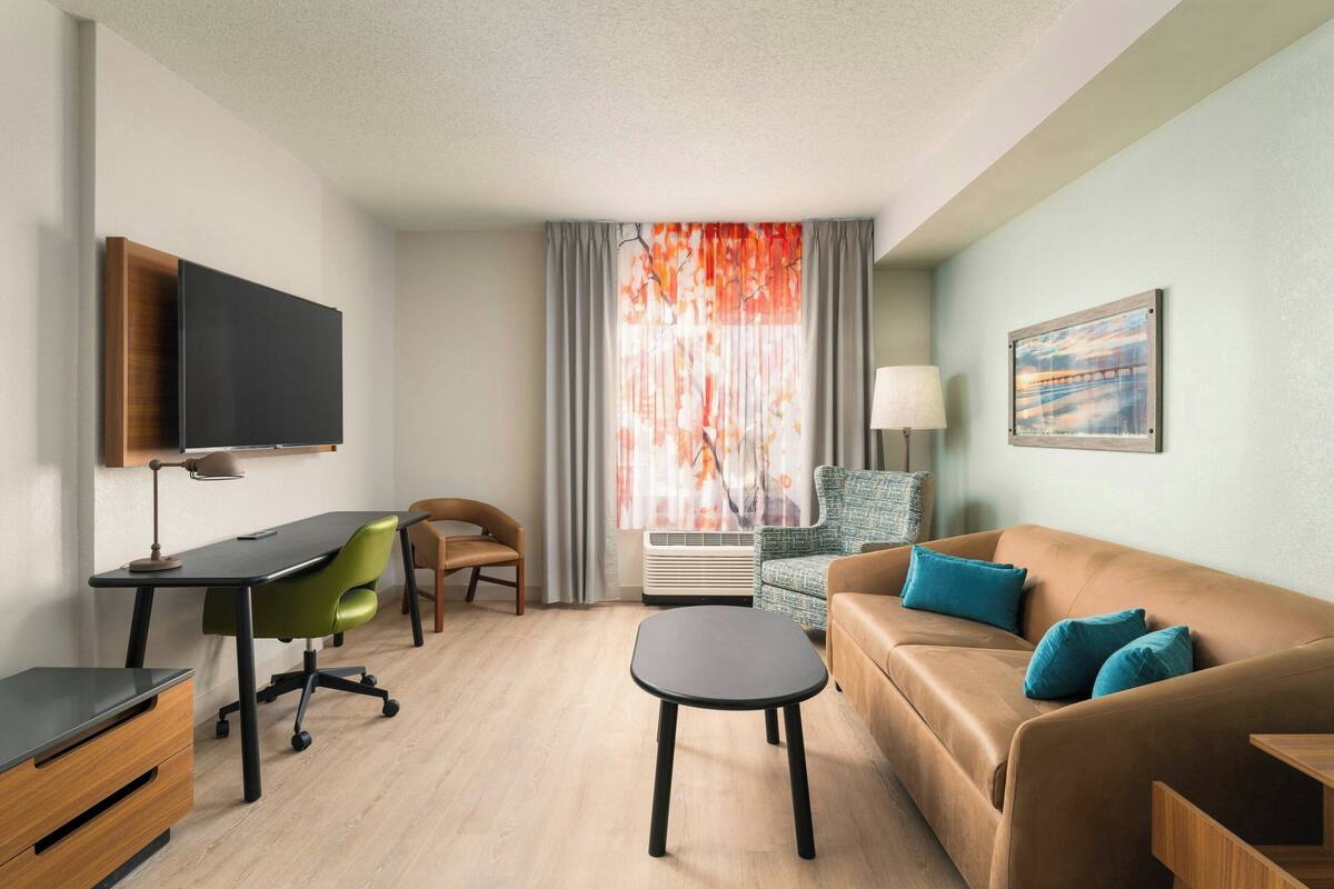 Una habitación de hotel moderna con escritorio, una silla verde, un sofá marrón y un televisor instalado en la pared.