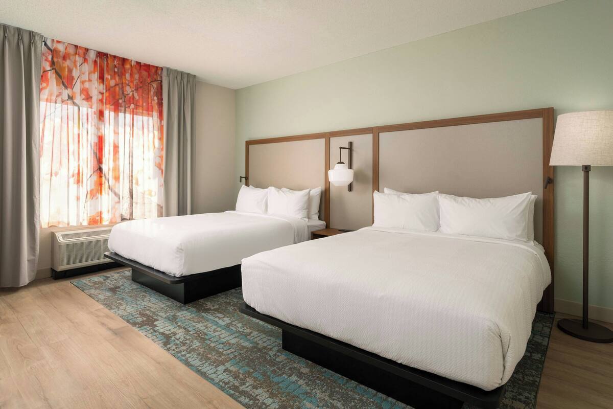 Habitación de hotel con dos camas, un ventanal con cortinas, una lámpara de pie y un calefactor portátil.