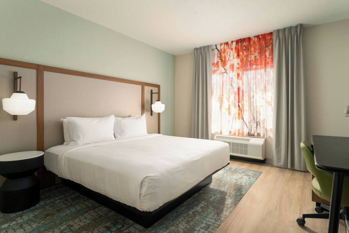 Habitación de hotel con cama, escritorio, silla, mesita redonda, ventana con cortinas y aire acondicionado de pared.