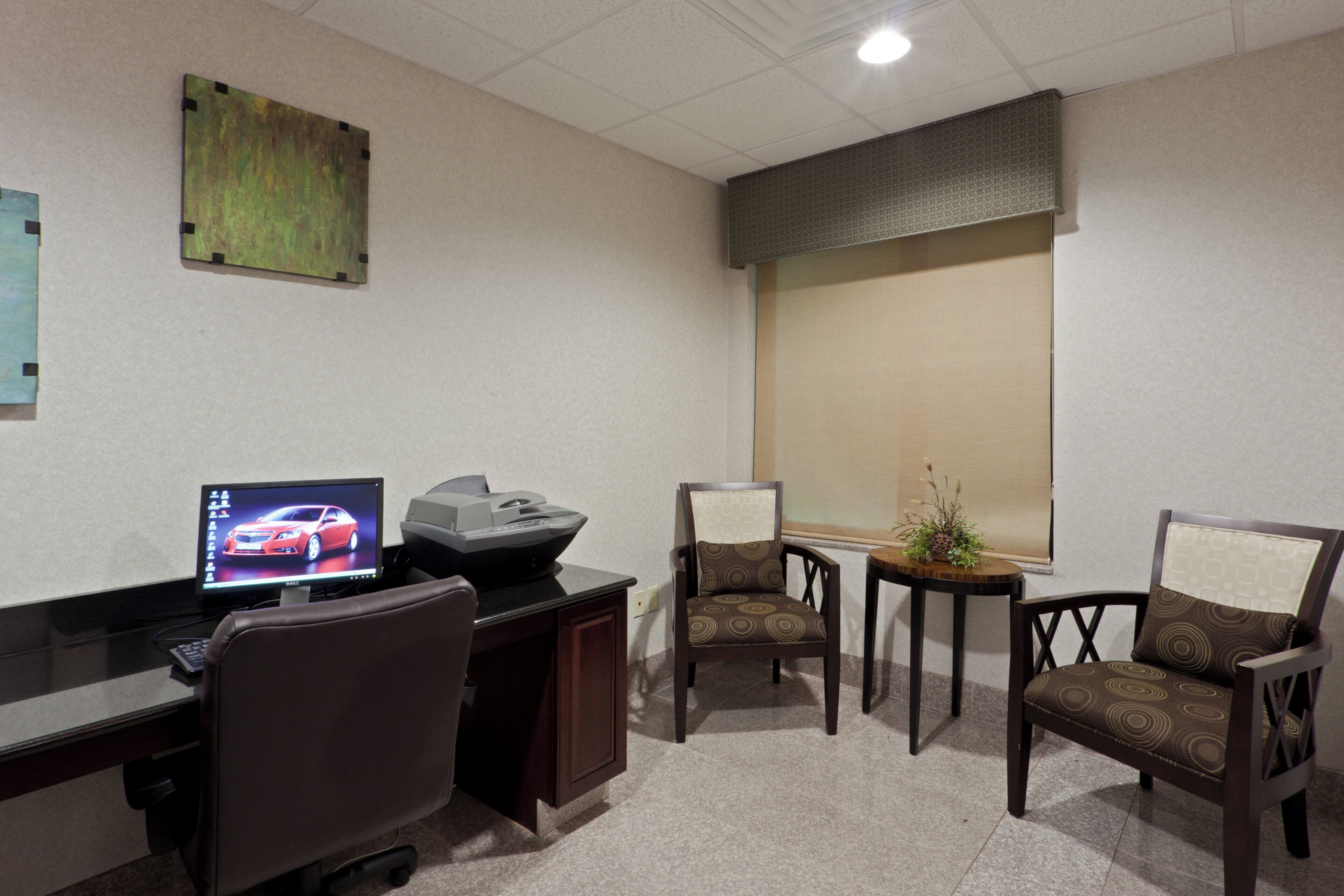 A conference room with a desk, computer, printer, two chairs, a small table with a plant, and a window with blinds.