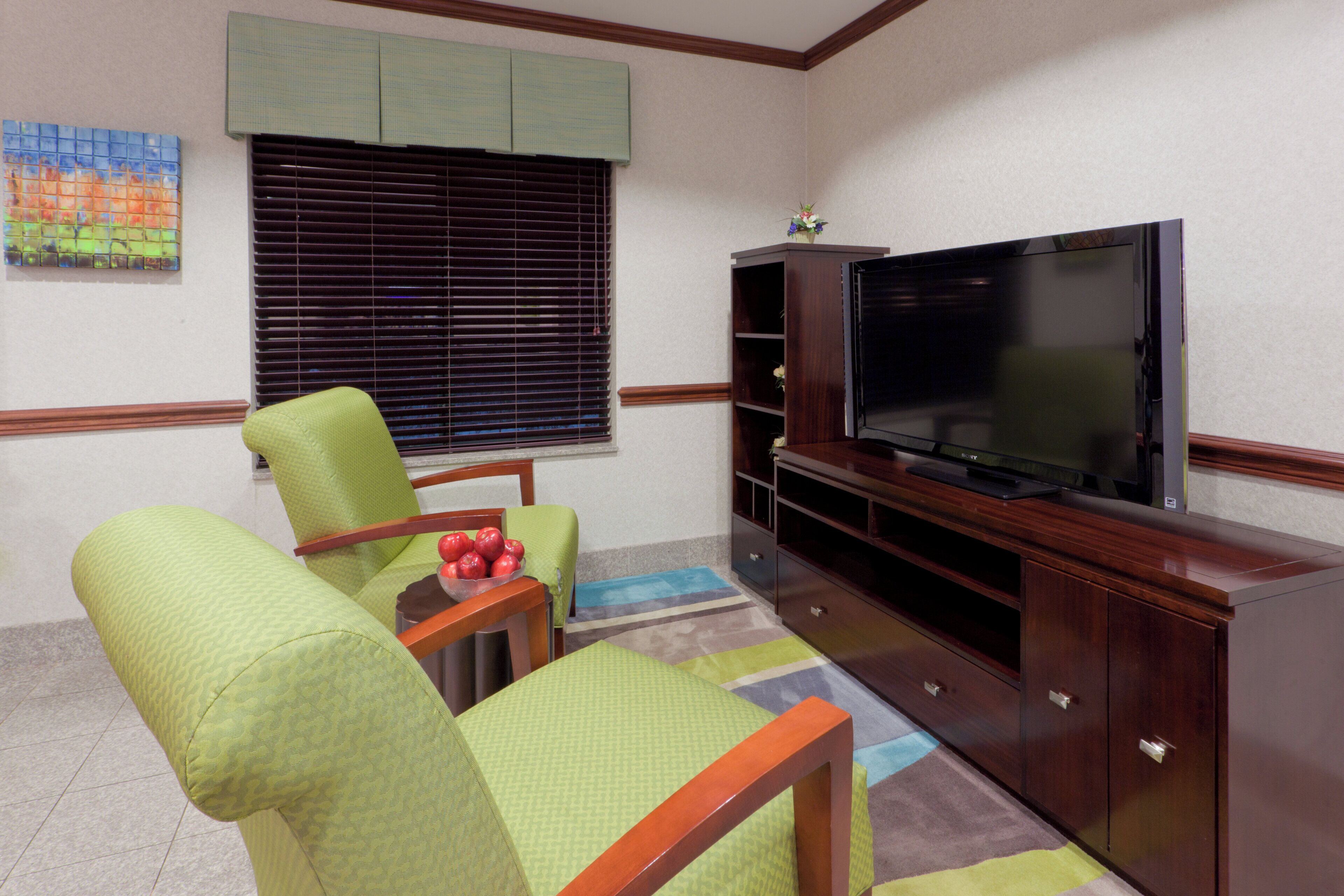 Una sala de estar con televisor de pantalla plana, sillones tapizados en verde, una mesita con manzanas y una ventana con persianas.