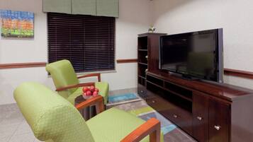 Una sala de estar con televisor de pantalla plana, sillones tapizados en verde, una mesita con manzanas y una ventana con persianas.