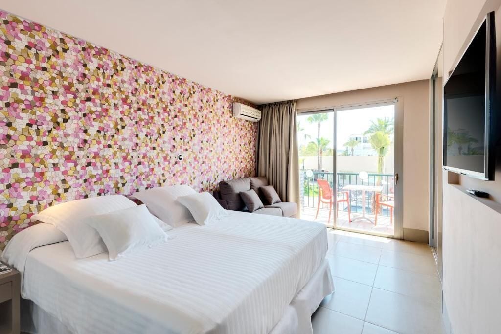 A hotel room with two beds, a balcony with a table and chairs, and a TV mounted on the wall.