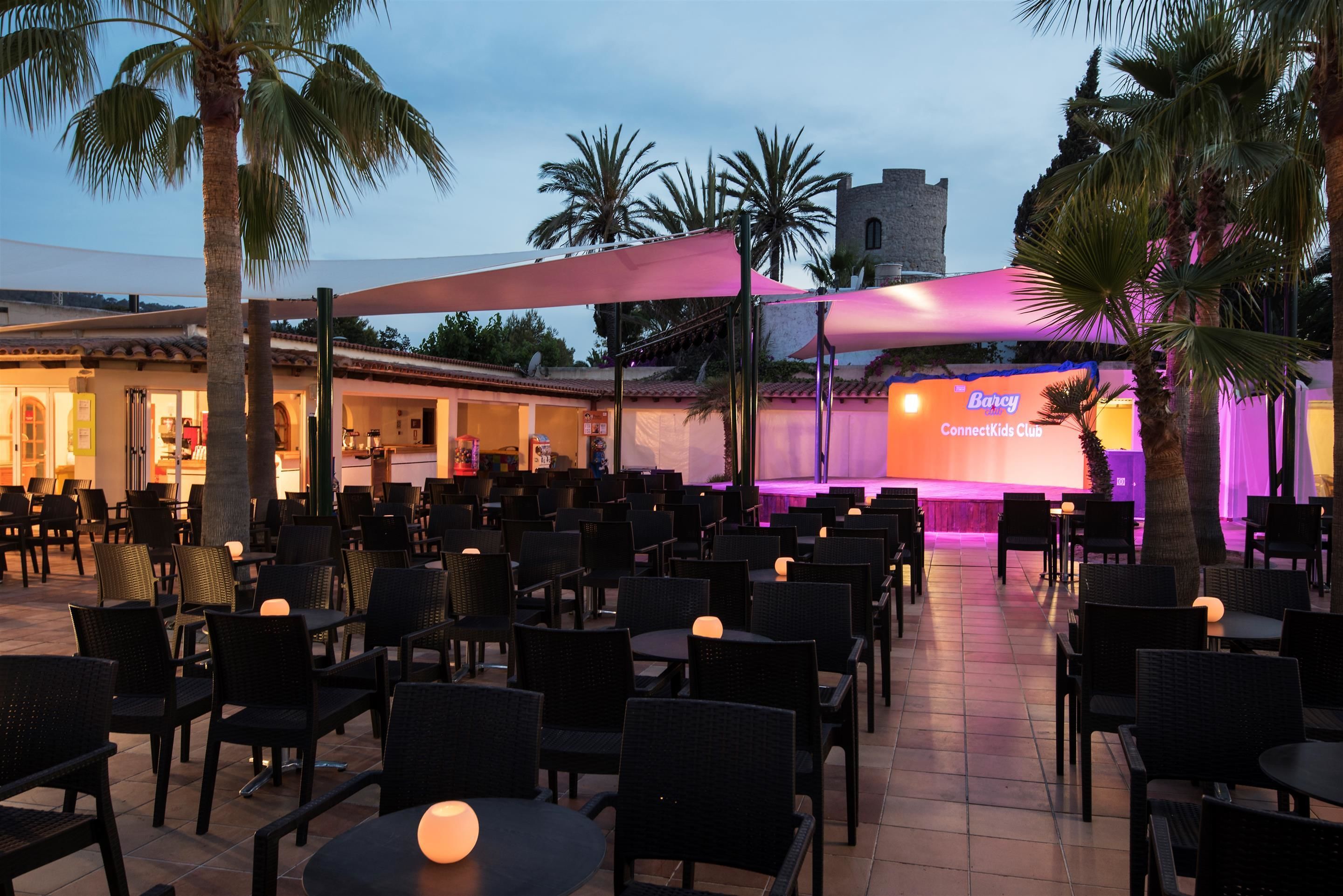 An outdoor seating area with tables and chairs, a stage with a banner, palm trees, and a building in the background.
