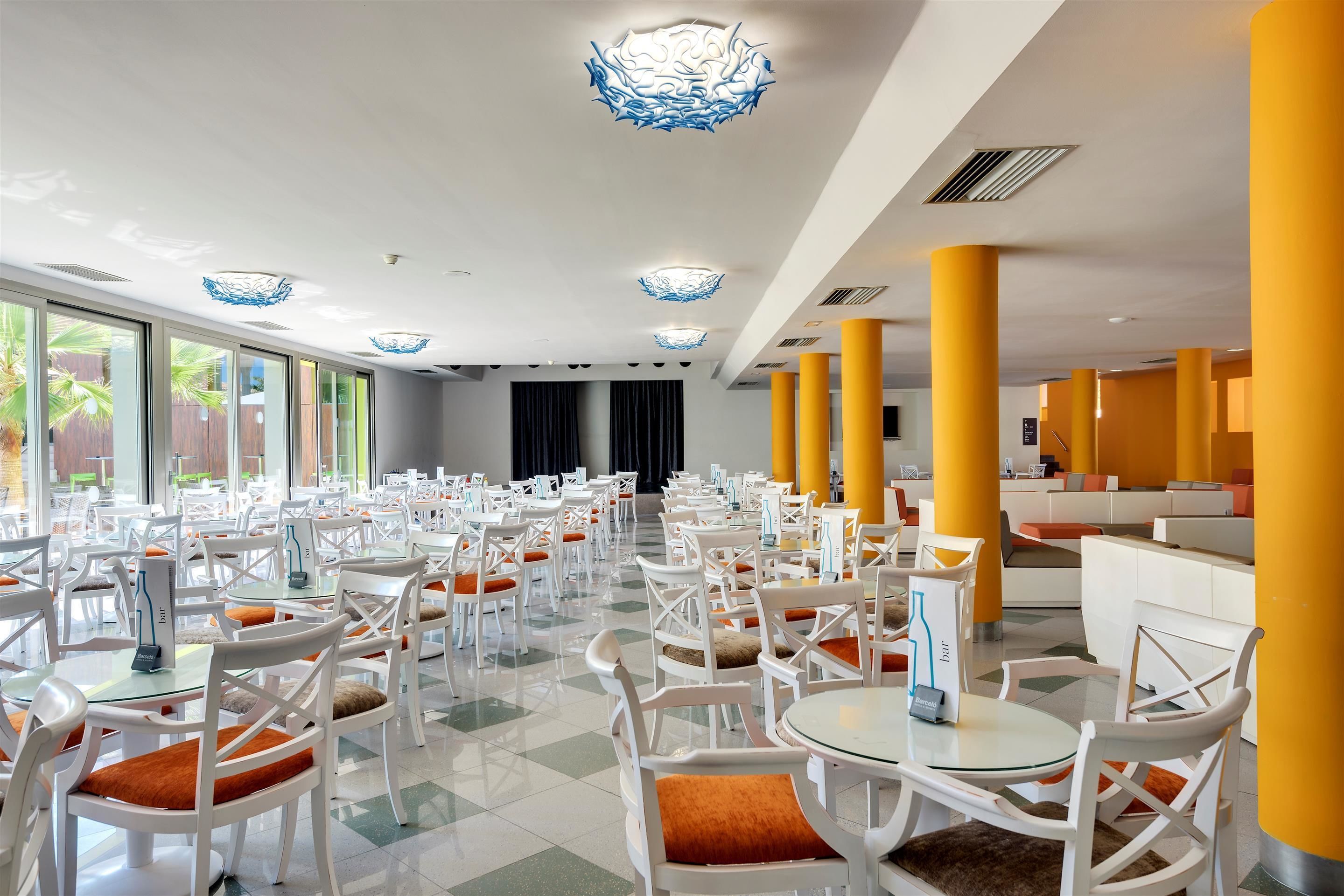 Un amplio comedor interior con mesas y sillas blancas, pilares amarillos e iluminación decorativa en el techo.