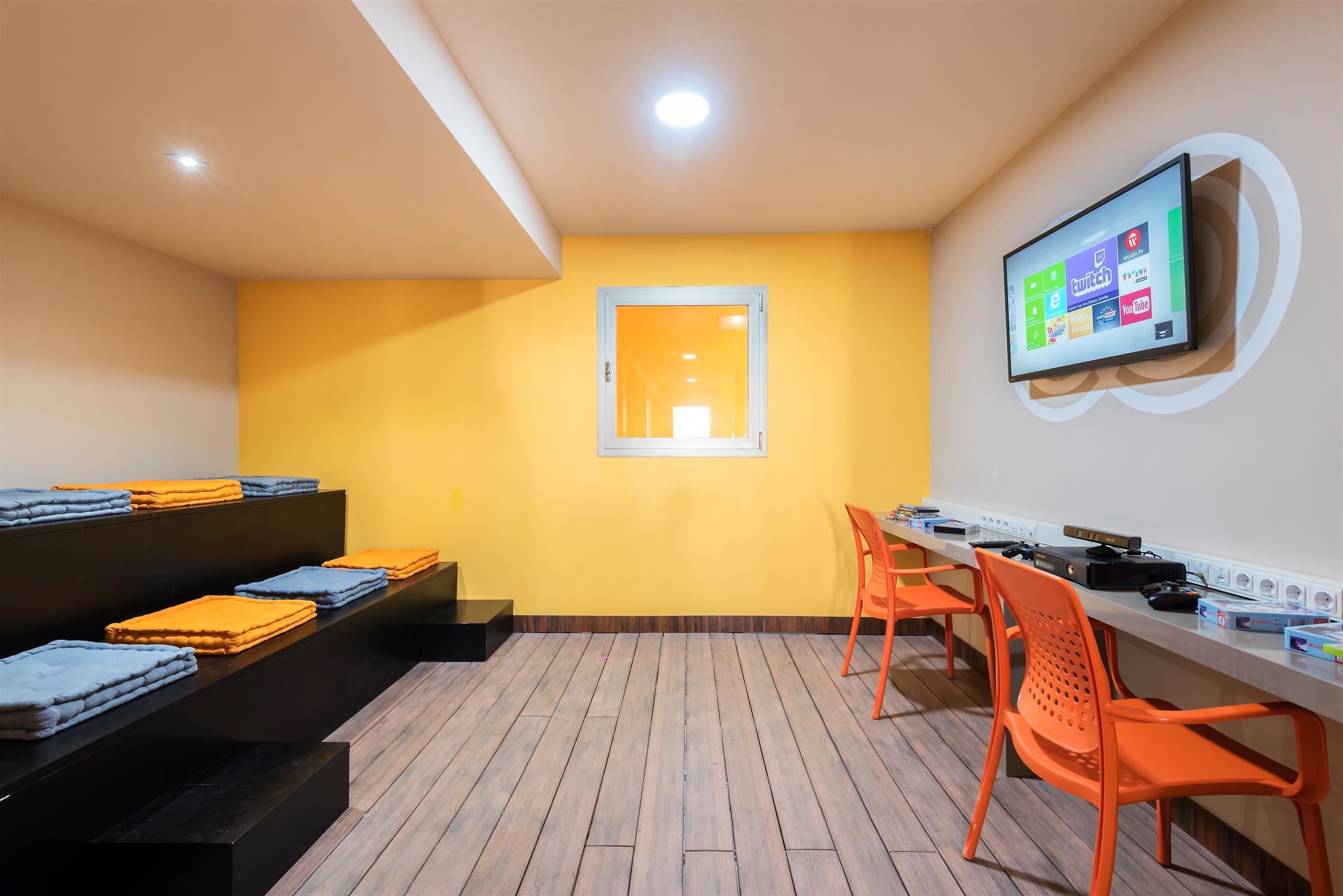 Una habitación con pared amarilla, ventana, televisor de pantalla plana montado en la pared, escritorio con computadora y sillas naranjas.