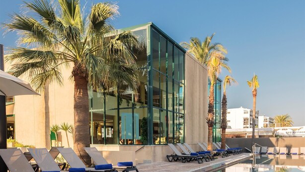 A pool area with lounge chairs, palm trees, and a modern building with large windows.